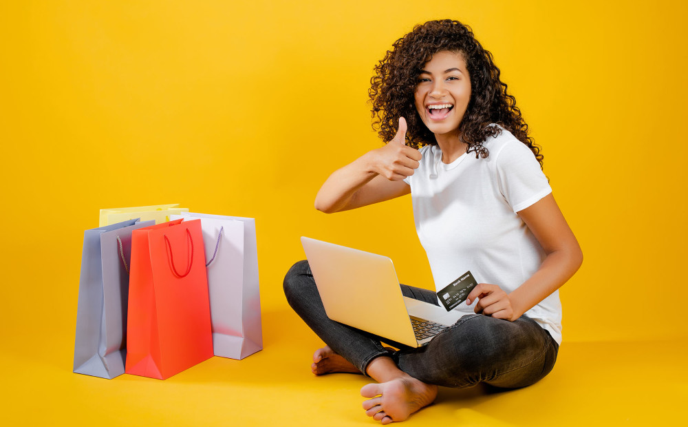 happy girl with colorful shopping bags sitting with laptop and credit card isolated over yellow