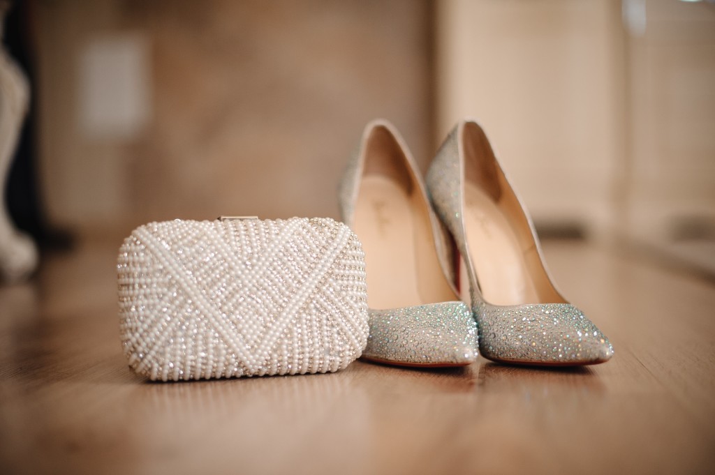 graceful shoes of the bride, studded with shiny beads, stand on the beige floor next to a small white clutch decorated with beads